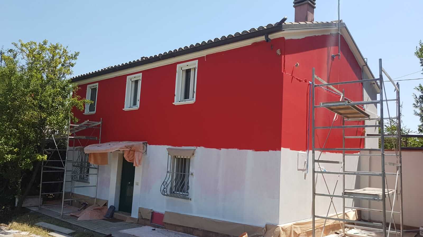 Casa con facciata rossa in fase di tinteggiatura, ponteggi sui due lati e teli di protezione a terra