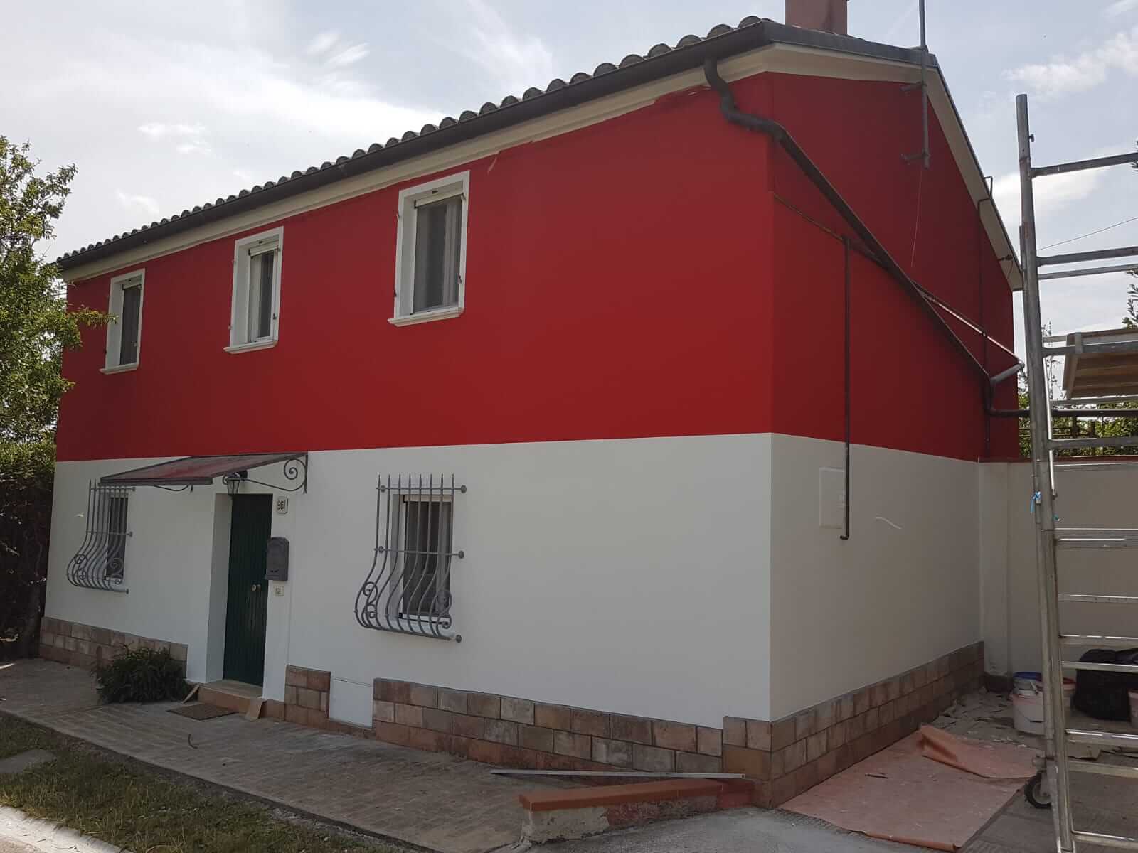 Vista d'angolo di una casa a due piani con fascia superiore rossa e zoccolatura bianca, ponteggio ancora montato sul lato
