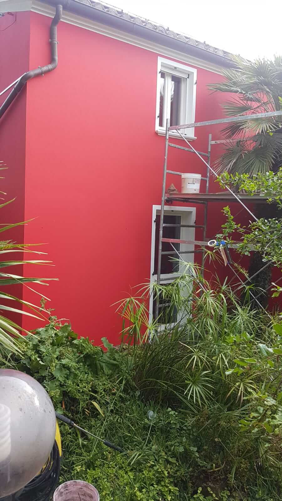 Parete laterale tinteggiata di rosso acceso con ponteggio montato e vegetazione in primo piano