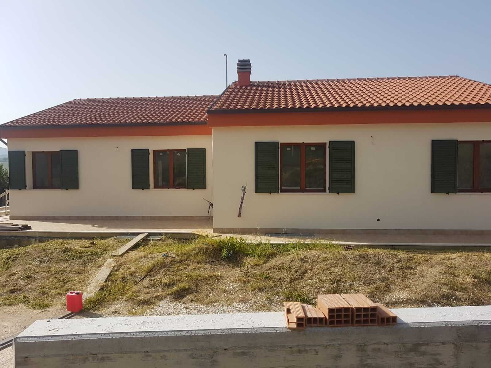 Prospetto frontale completo di una villa a un piano con pareti crema, fascia arancione sotto il tetto e persiane verdi